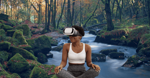 A woman sits cross-legged with a VR headset on in front of a peaceful stream in the forest.